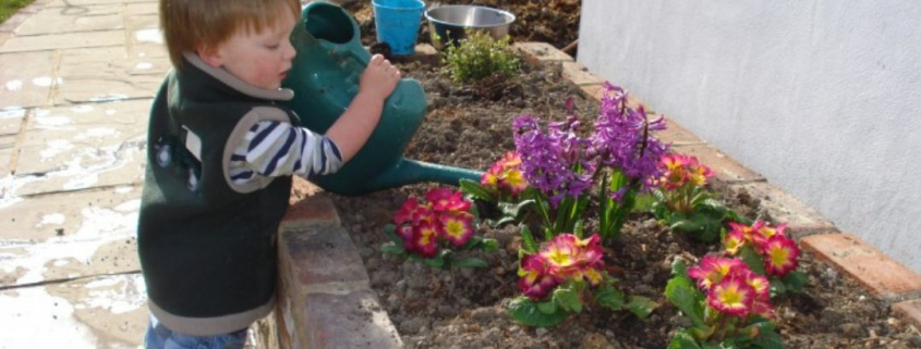 a young child in a jumper and wellies watering a flower bed with a plastic watering can