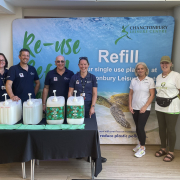 Six people standing in front of a large “Refill” banner at Chanctonbury Leisure Centre, smiling beside a display of refillable household product dispensers. Three people wear navy uniforms, and two wear Sussex Green Living T-shirts. The backdrop features a sea turtle and the words “Re-use, Refill, Reduce.”
