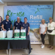 Six people standing in front of a large “Refill” banner at Chanctonbury Leisure Centre, smiling beside a display of refillable household product dispensers. Three people wear navy uniforms, and two wear Sussex Green Living T-shirts. The backdrop features a sea turtle and the words “Re-use, Refill, Reduce.”