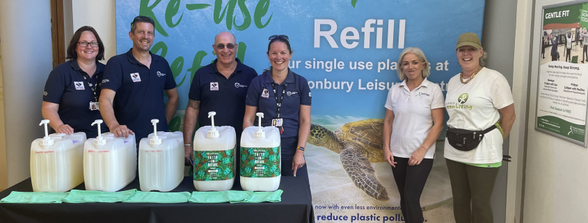 Six people standing in front of a large “Refill” banner at Chanctonbury Leisure Centre, smiling beside a display of refillable household product dispensers. Three people wear navy uniforms, and two wear Sussex Green Living T-shirts. The backdrop features a sea turtle and the words “Re-use, Refill, Reduce.”
